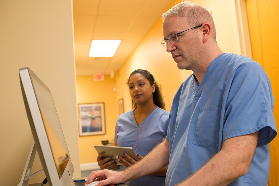 male doctor and female assistant in blue scrubs looking at desktop computer screen