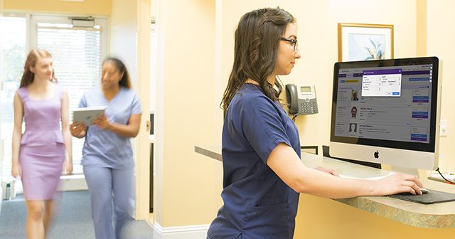 Woman in navy scrubs using dermatology practice management system and patient and medical assistant in background