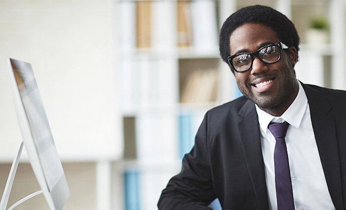 Person smiling, wearing glasses and a suit next to a desktop computer