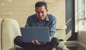 smiling male patient sitting on couch at home using laptop