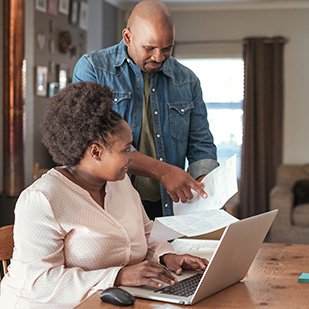 male and female smiling, pointing at paperwork and working on a laptop