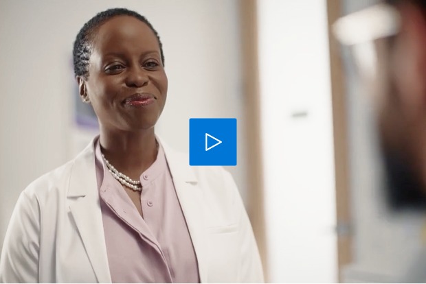 Video thumbnail showing a smiling female doctor in a white coat, light purple blouse, and pearl necklace, standing in a bright medical office, with a blue play button icon in the center.