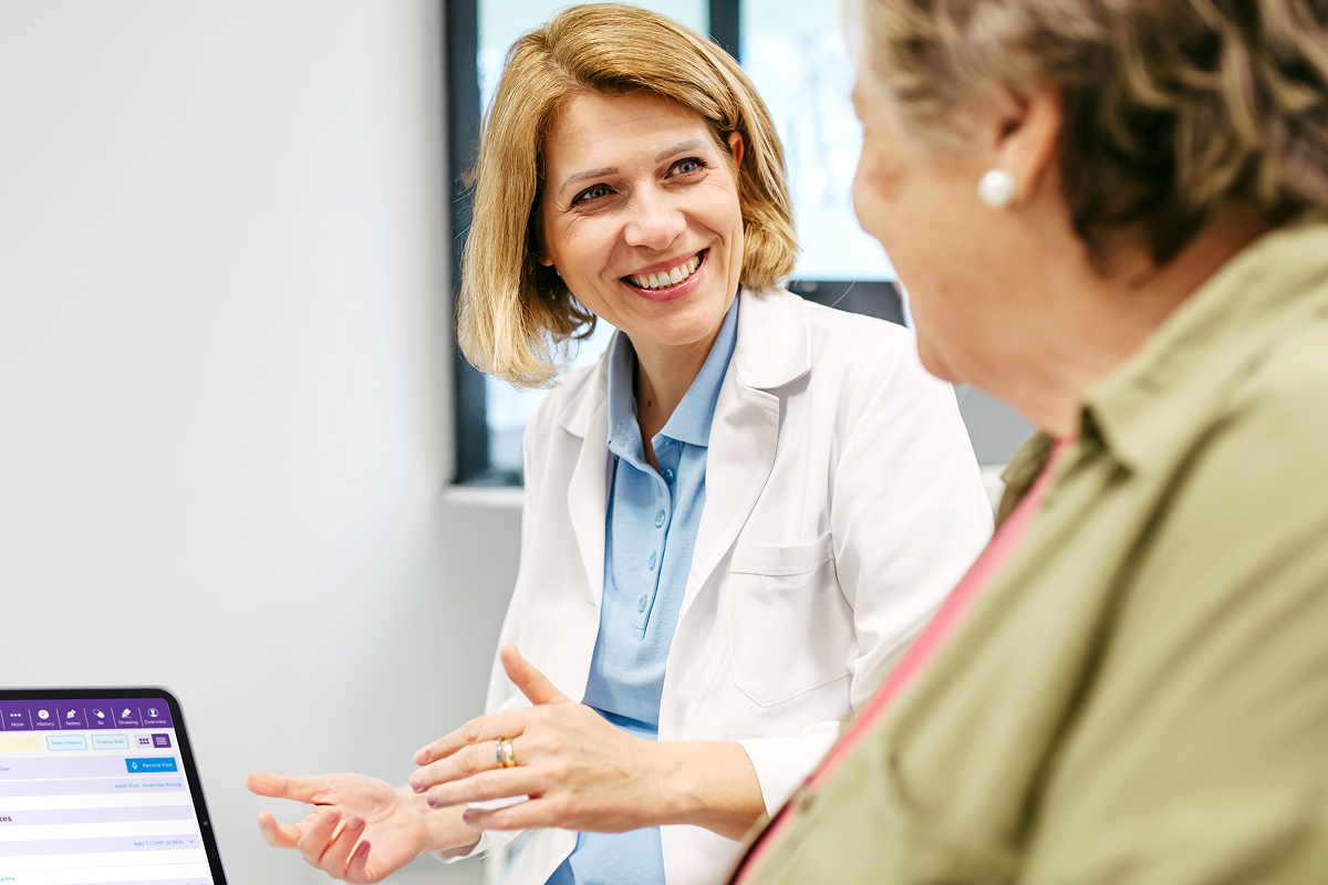 A smiling physician in a white coat consults with a patient, gesturing toward a tablet running the mobile EHR.