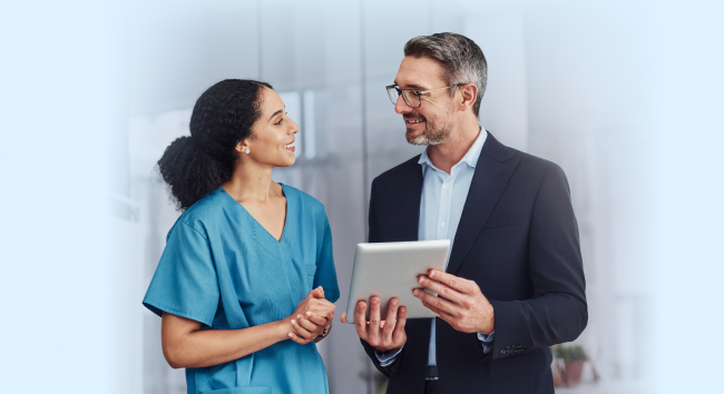 A smiling female healthcare professional in teal scrubs and a smiling male executive in a suit and glasses conversing while looking at a tablet together in a bright, modern clinical setting.