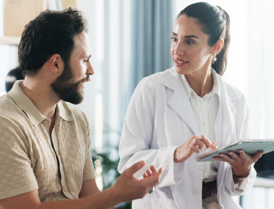 A female doctor in a white lab coat holds a tablet and smiles while having a conversation with a male patient in a brightly lit consultation room.