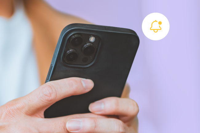 Un primer plano de las manos de una persona que sostienen un smartphone negro, acompañado de un círculo blanco flotante que contiene un icono amarillo con forma de campana de notificación.