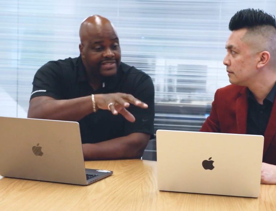 Two men sitting at a desk with open laptops, collaborating in an office setting. One man in a black polo shirt is gesturing and speaking, while the other man in a red jacket listens attentively.