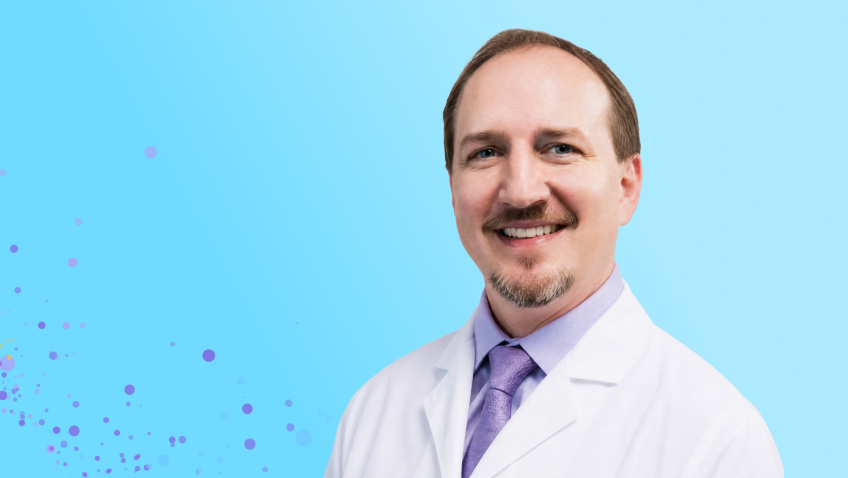 Médico sonriente con bata blanca, camisa morada y corbata, sobre un fondo azul claro