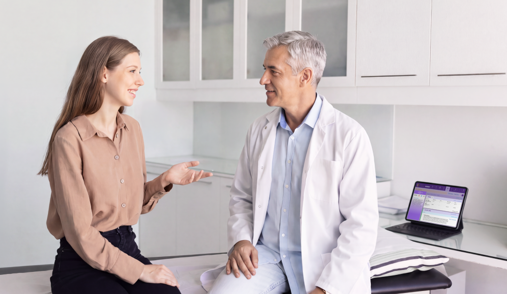 Una paciente hablando con un médico sentado en una camilla de consulta en una clínica, con una tableta cerca