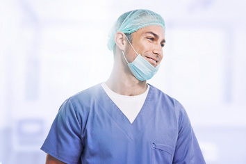 Un profesional sanitario de sexo masculino, sonriente, con bata azul, que lleva un gorro quirúrgico y una mascarilla quirúrgica bajada hasta debajo de la barbilla.